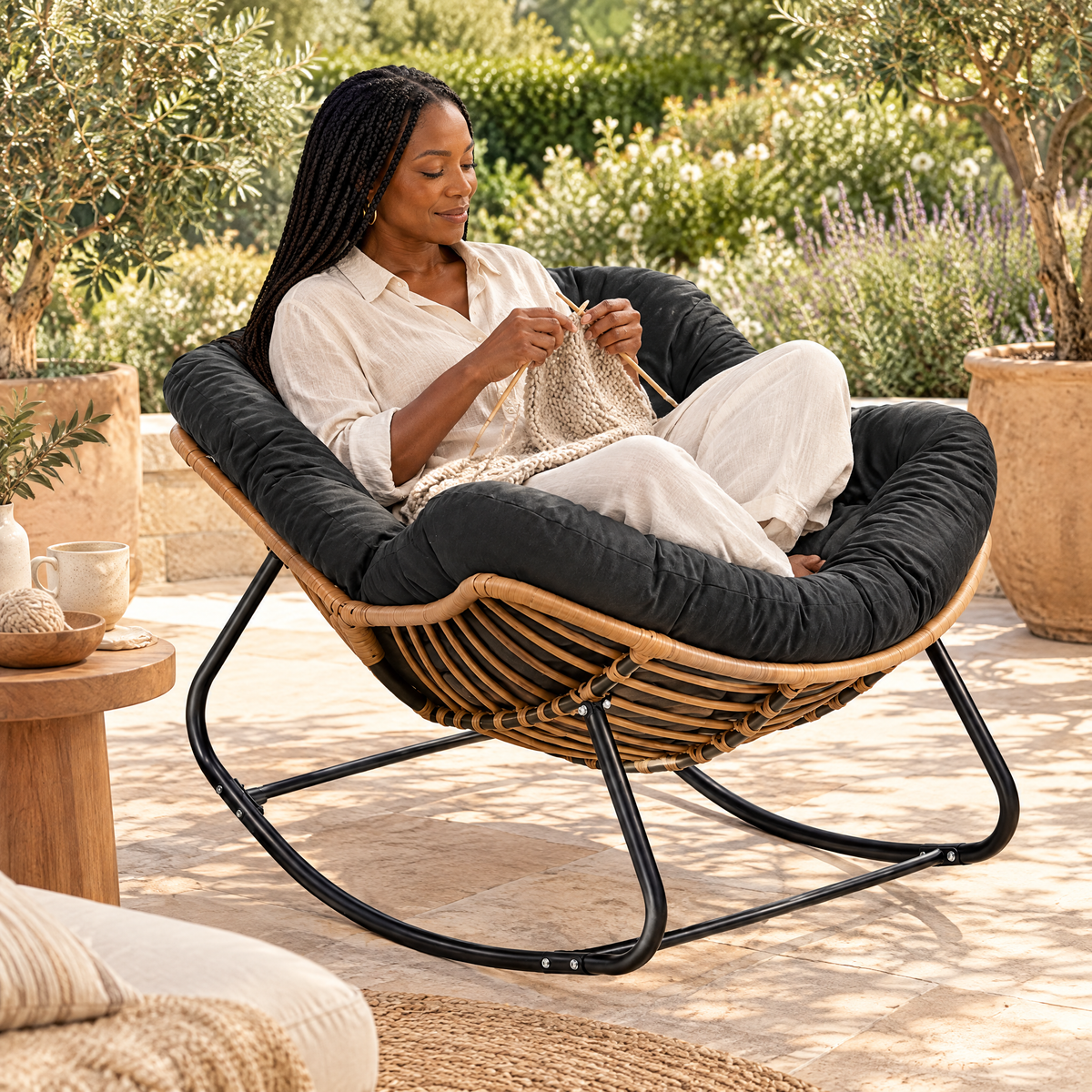 A Black woman with long box braids in her mid 40s, knitting, in The Cabana indoor-outdoor rattan rocker in a sunny stone patio