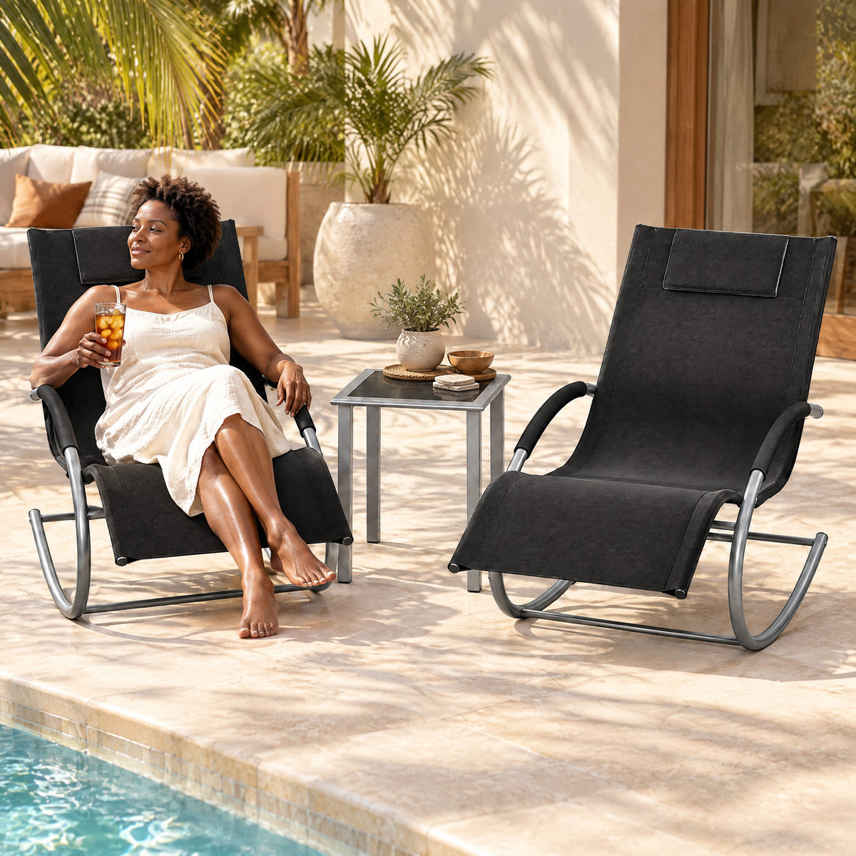 A Black woman with short natural curls in her mid 40s, holding a mug of tea and looking out the window, in The Lido set: 2 rocking sun loungers + side table in a poolside deck